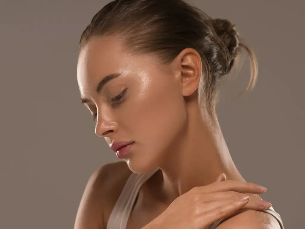 Close-up of a woman with smooth, glowing skin, hair tied in a neat bun, gently touching her shoulder while looking down against a soft neutral background.