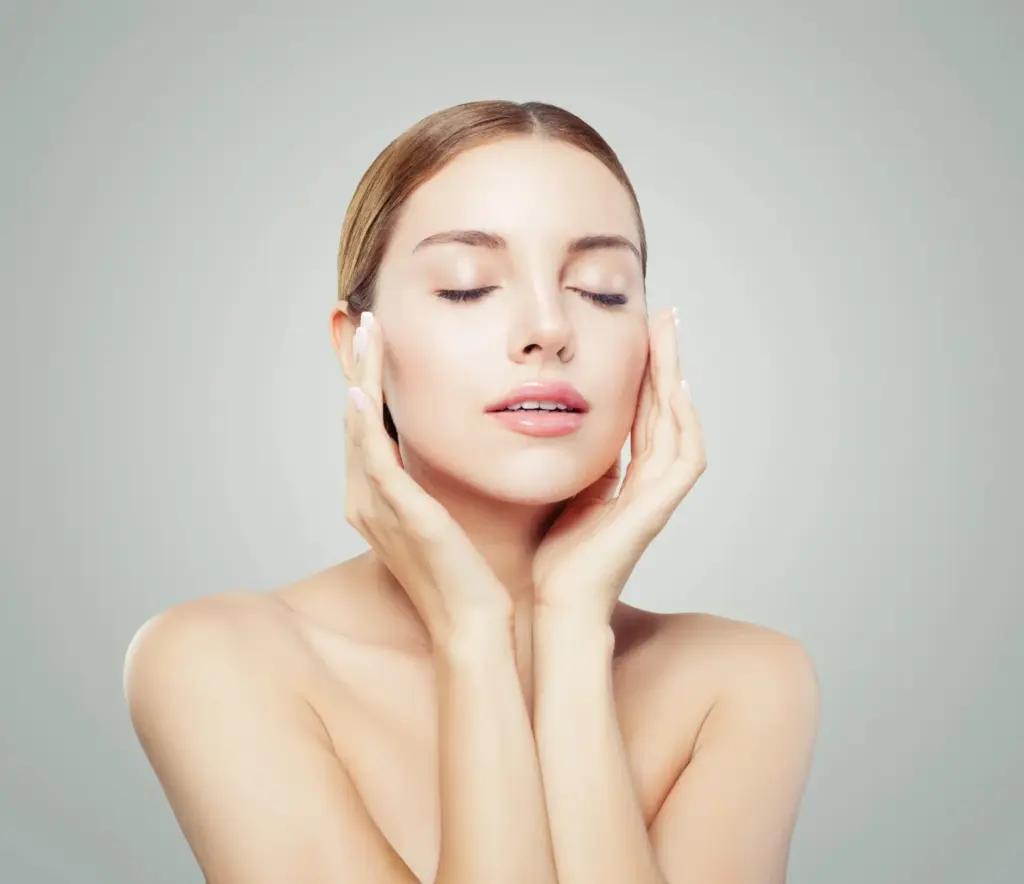 Portrait of a woman with smooth, radiant skin, eyes closed, gently touching her face with both hands against a soft neutral background, suggesting skincare or cosmetic treatment results.